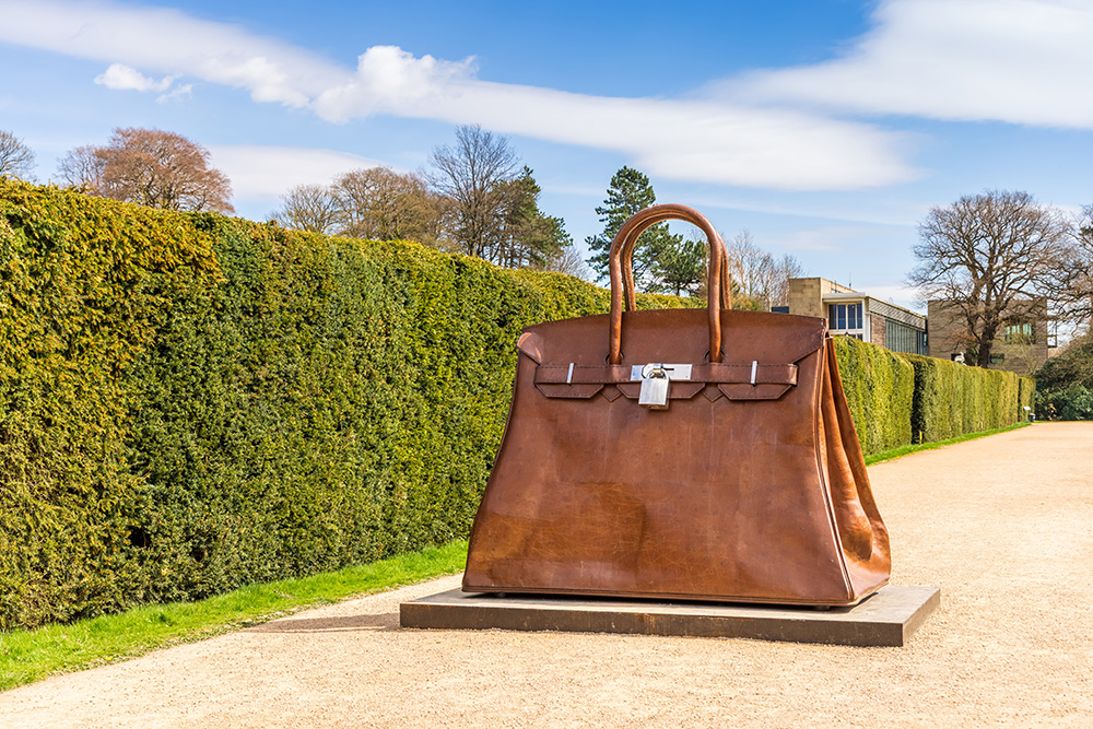 A large-scale bronze sculpture of an Hermes Birkin bag in an outdoor setting, with a lush hedge and clear sky in the background.